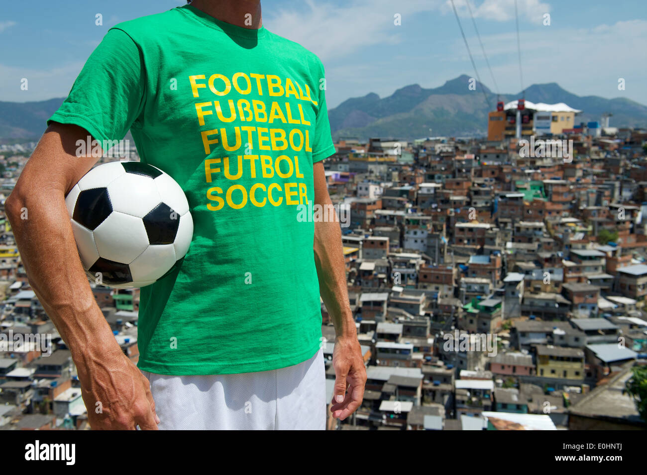 Brazilian man standing in international football t-shirt holding soccer ball in front of favela slum background Rio de Janeiro Stock Photo