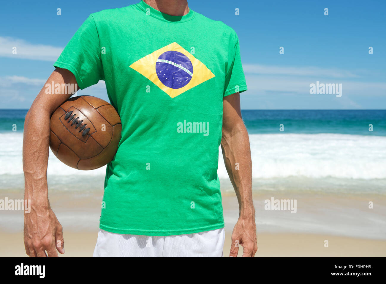 Brazilian soccer player in Brazil flag shirt holding vintage football on tropical Ipanema Beach Rio de Janeiro Stock Photo