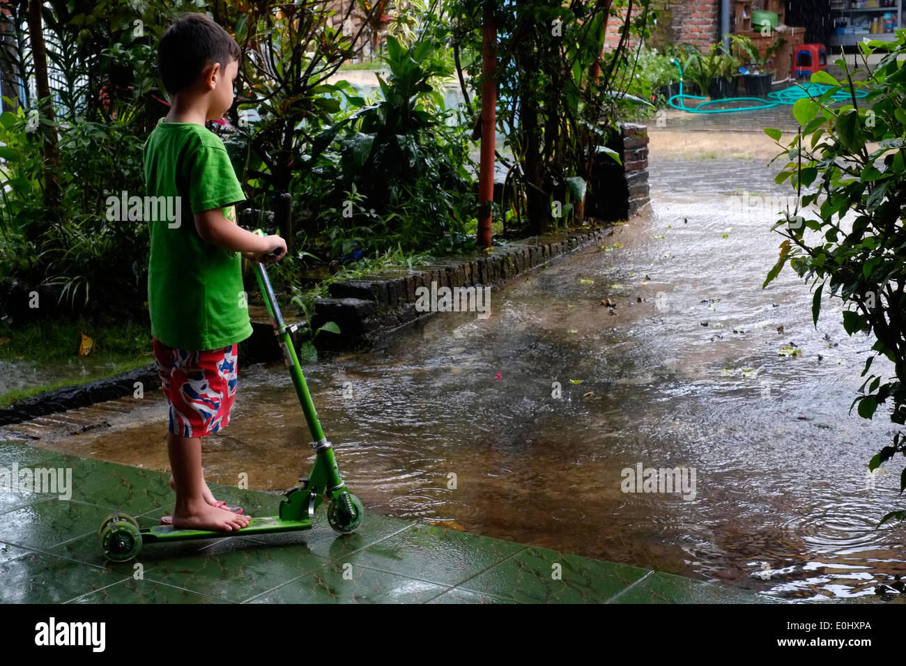 little boy with his scooter looking wistfully at the pouring rain as he waits to go out and play in rural village indonesia Stock Photo