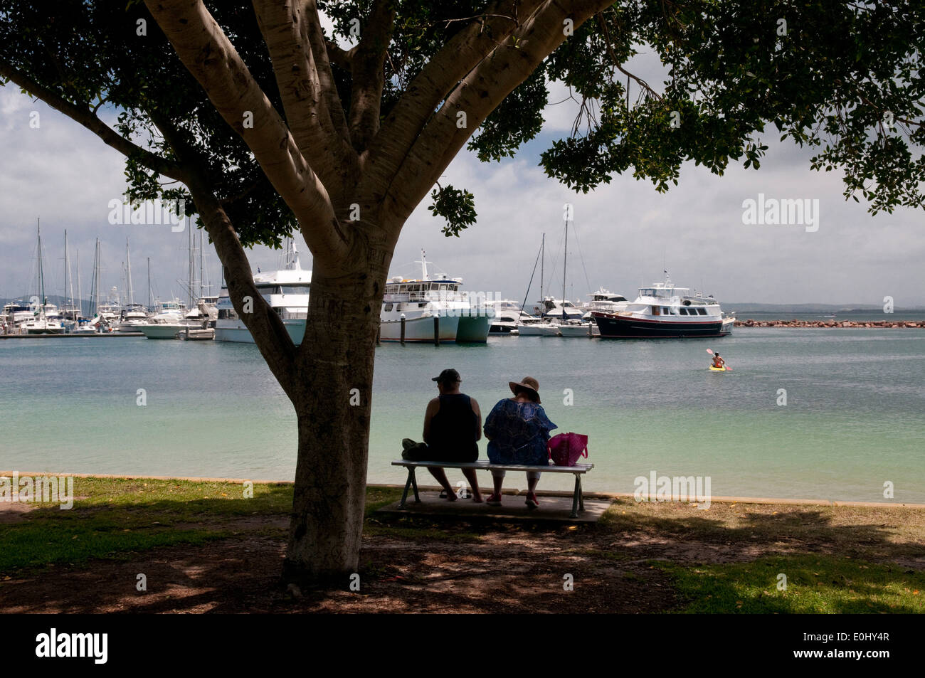 A couple sit in the shade of a tree overlooking the marina at Nelson Bay, Port Stephens, New South Wales, Australia. Stock Photo