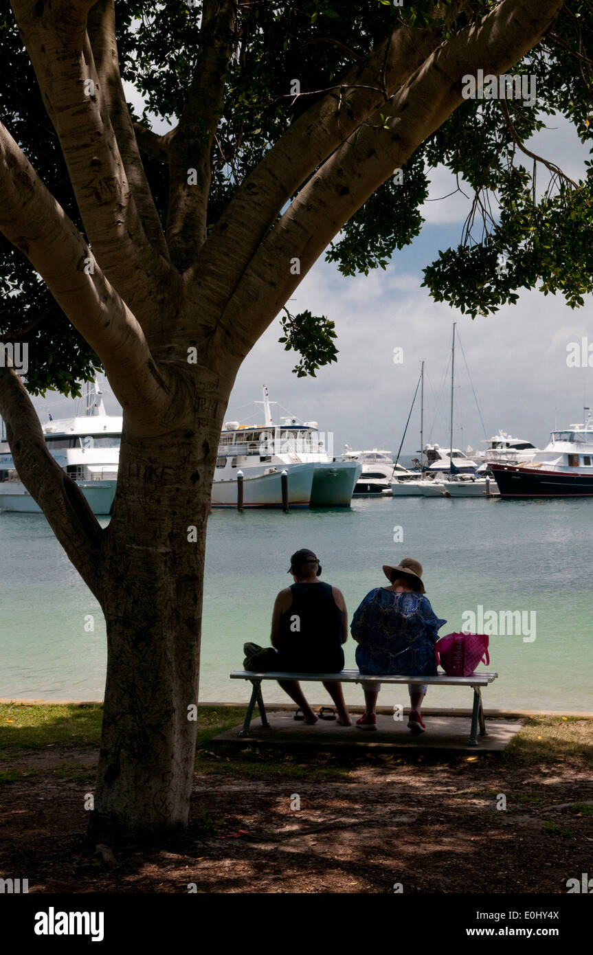 A couple sit in the shade of a tree overlooking the marina at Nelson Bay, Port Stephens, New South Wales, Australia. Stock Photo