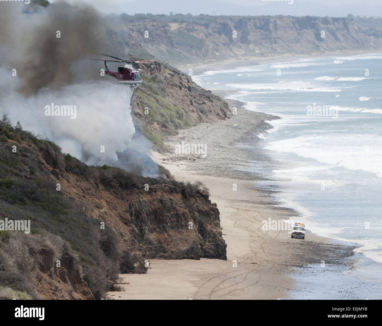 San Onofre, California, USA. 13th May, 2013. A California State Lifeguard jeep makes its way past a flames burning along a bluff area just above the water's edge near Trail One at San Onofre State Campground on Wednesday morning as a waterdropping helicopter drops a load of ocean water on the fire. Multiple Fire agencies, including Federal, State and County, responded to a wildfire on at the San Clemente Check Point that began when a truck on the northbound I-5 Freeway caught fire and passed to nearby vegetation on Wednesday morning. The Pendleton West Fire, had north and south bound traffic Stock Photo
