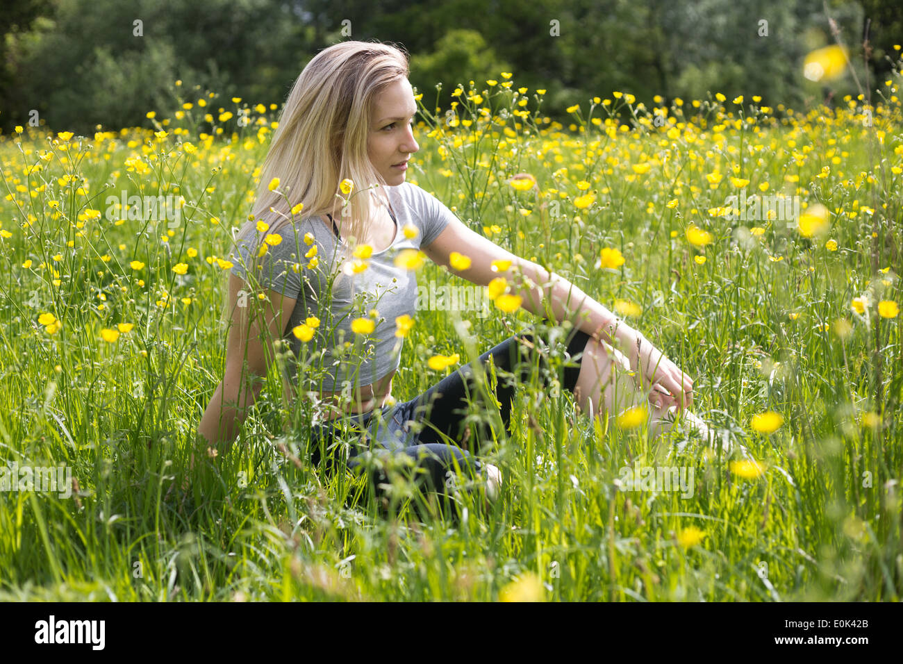 Blonde lady wearing black leggings and a grey crop top sitting in long grass with yellow flowers hampstead heath. Stock Photo