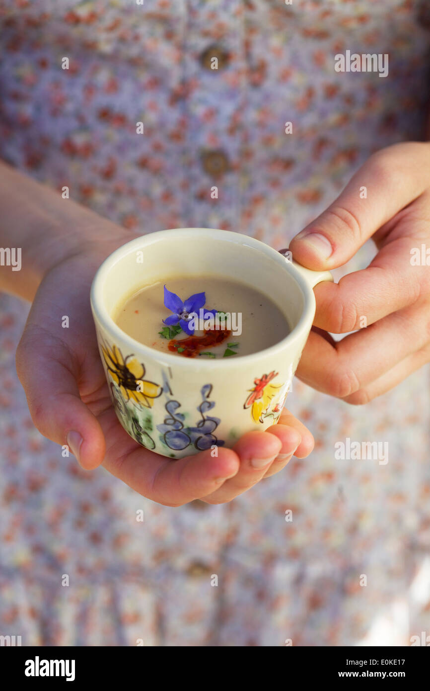 An close-up of a female's hands offering a small cup of soup garnished with chili oil, chopped green herb and a purple flower. Stock Photo