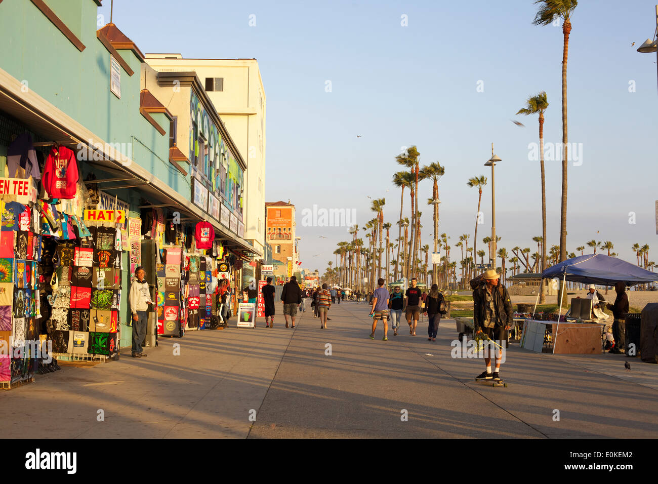 Shoppers walk and a young man skateboards down the sunny Venice Beach Boardwalk in Los Angeles, California. Stock Photo