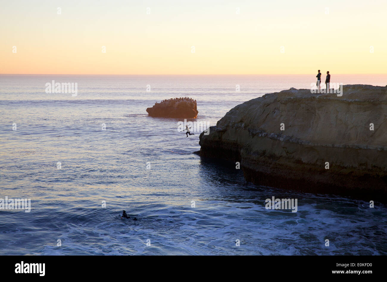 Onlookers watch on a cliff as a surfer jumps into the ocean at