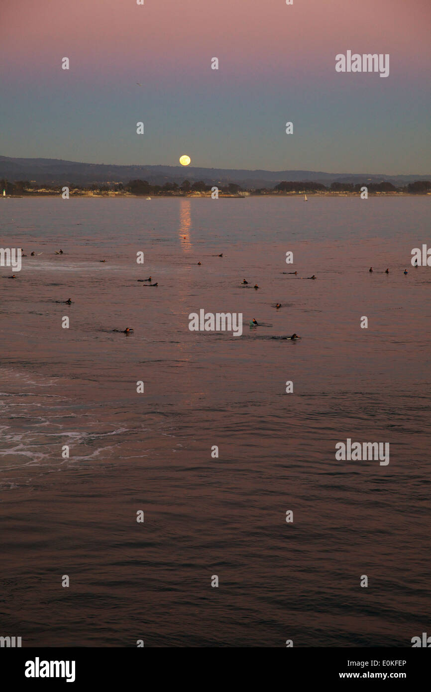 The full moon rises over the Pacific Ocean at Steamers, Santa Cruz while surfers wait to catch a wave in the foreground. Stock Photo