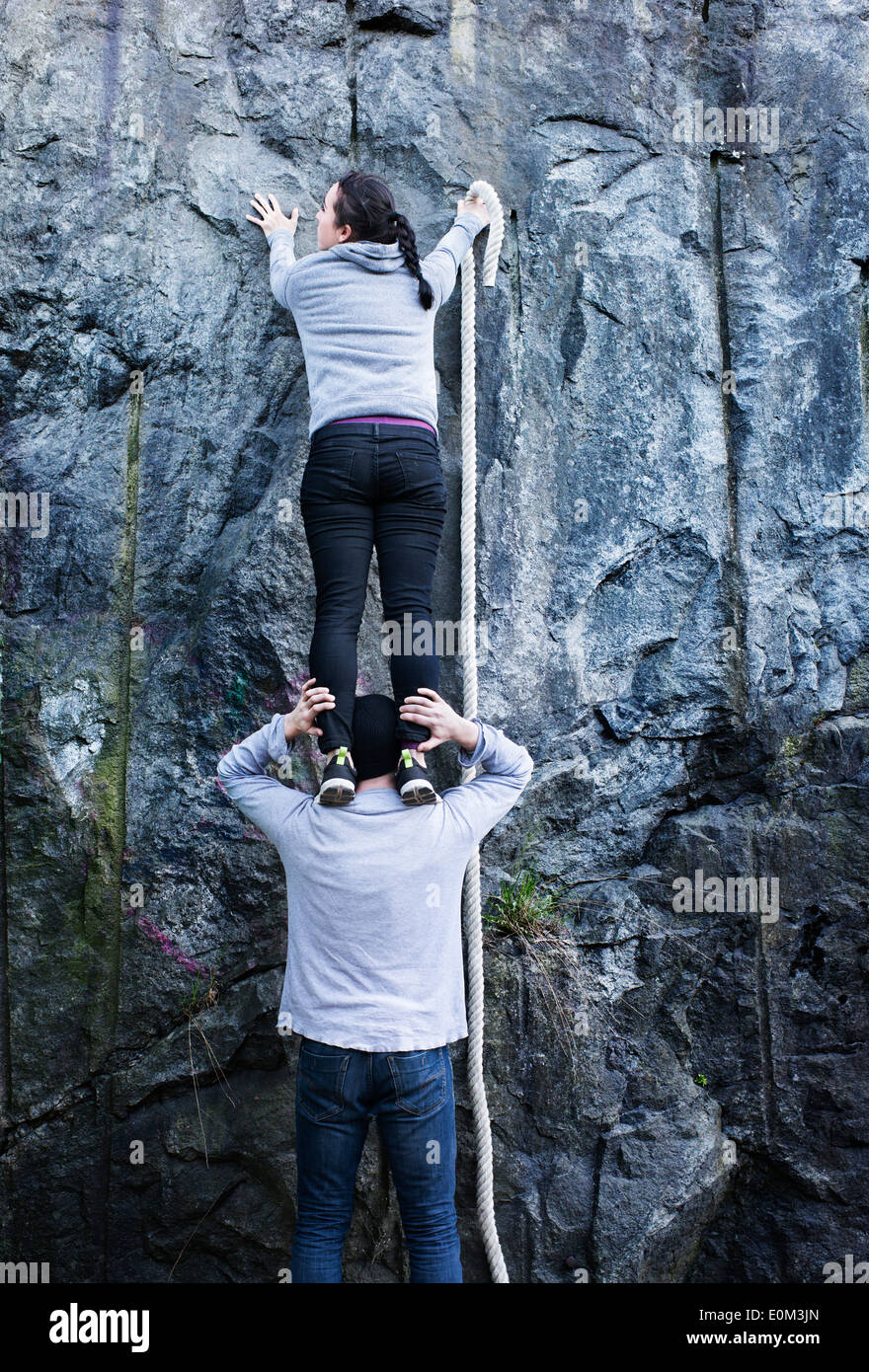 Woman standing on shoulders of man in front of mountain wall with rope in hand. Looking for a way to climb up. Stock Photo