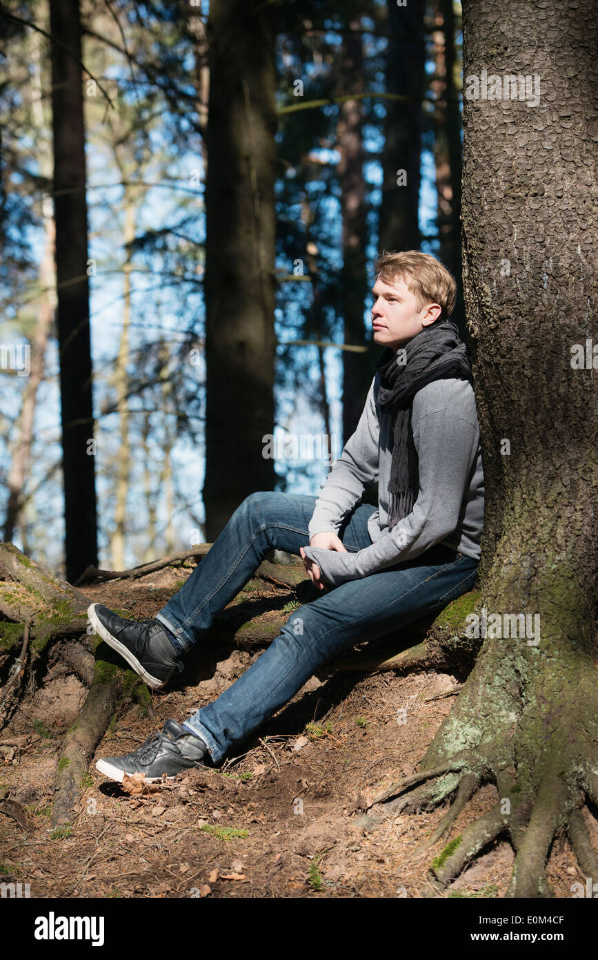 Young pensive man in nature resting by a tree in forest during a trek. Stock Photo