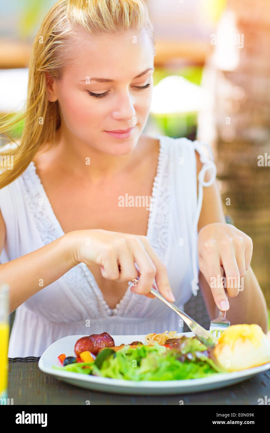 Portrait of attractive woman eating fresh salad and meat using knife and fork, having lunch in
