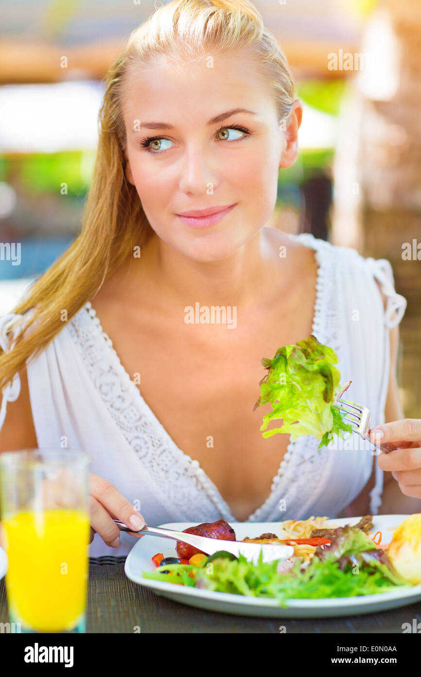 Portrait of attractive woman eating fresh green salad using knife and fork, having lunch in