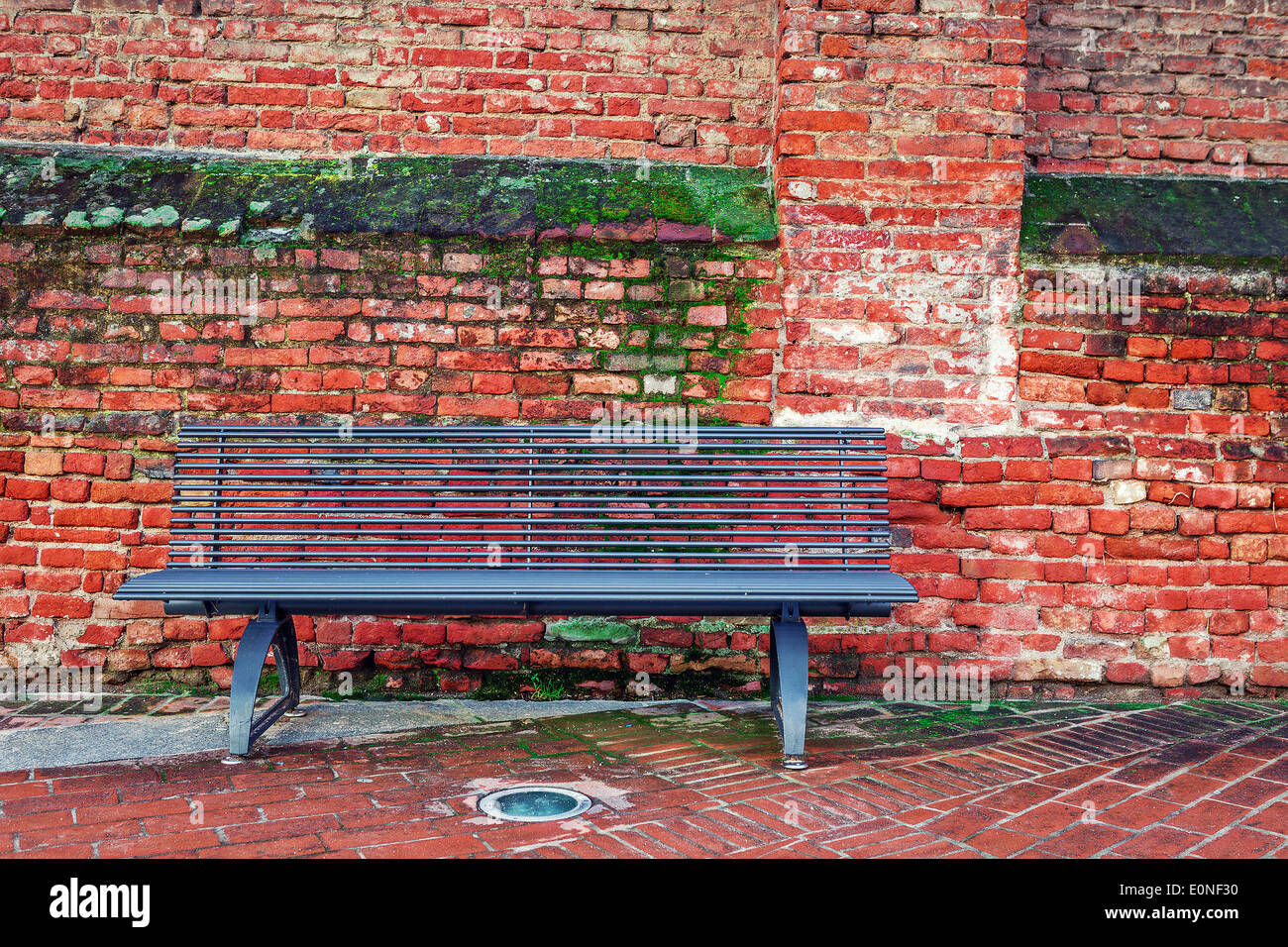 Metal bench on sidewalk against old red brick wall with green moss in Alba, Italy. Stock Photo