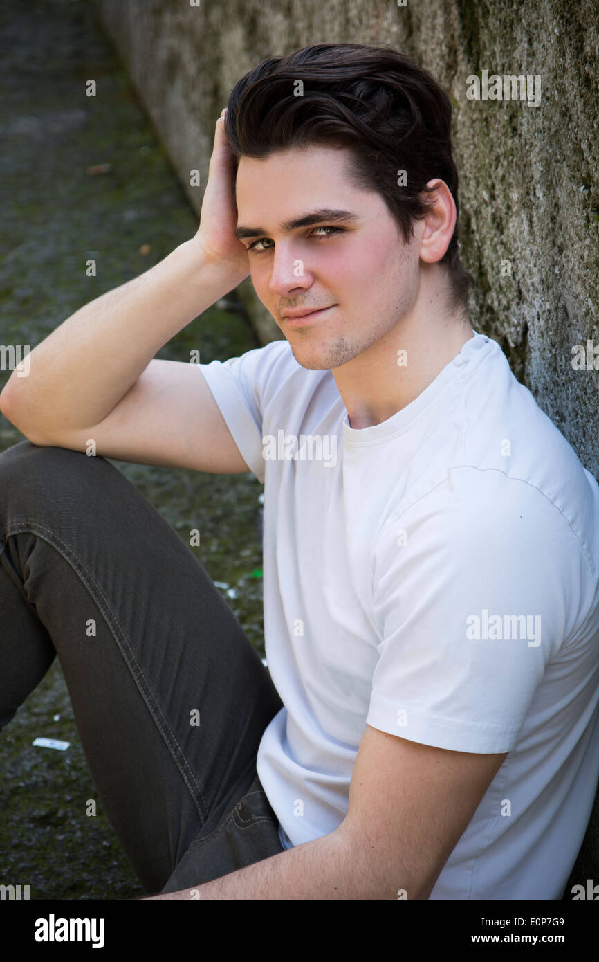 Attractive young man sitting on the ground against rock outdoors, looking at camera with a smile Stock Photo