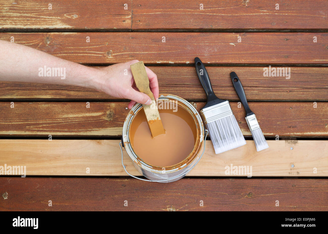 Horizontal photo of male hand stirring wood stain in can with stir stick with two paint brushes lying on a single new cedar wood Stock Photo
