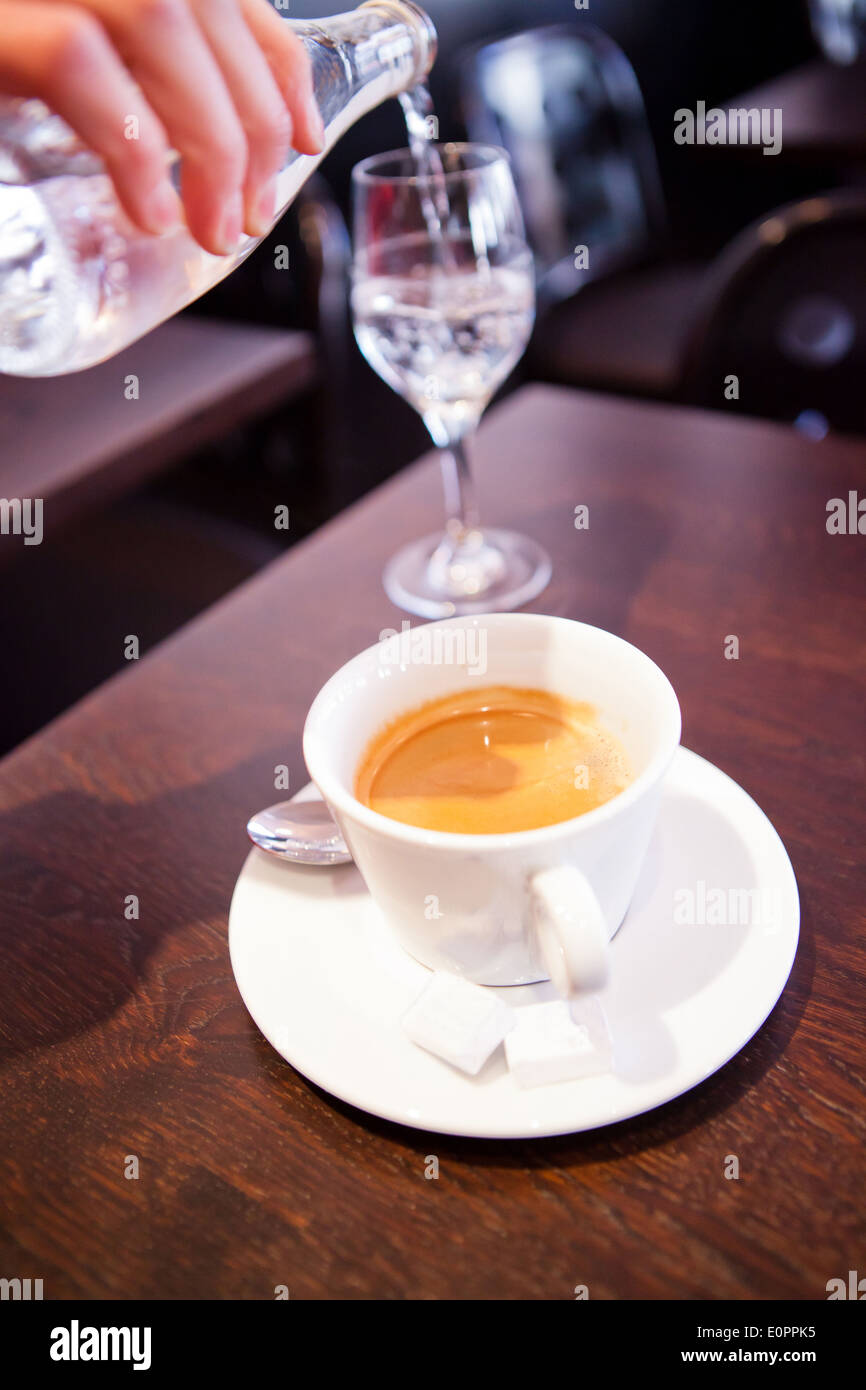 Closeup of cup of espresso coffee on wooden cafe table and pouring of water into glass from bottle in background Stock Photo