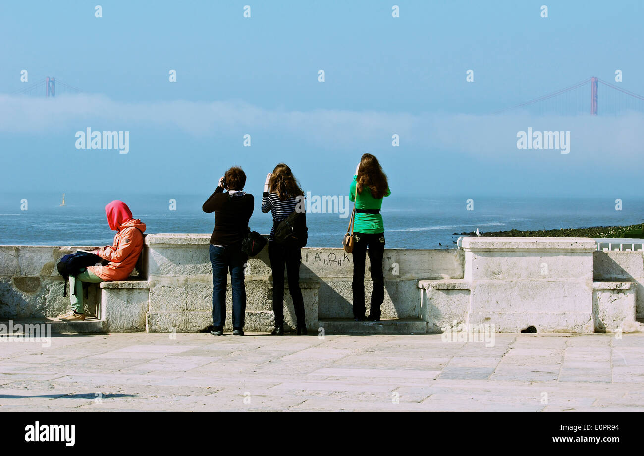 Woman sitting reading by river Tagus (Tejo) and 3 female tourists taking pictures Lisbon Portugal western Europe Stock Photo