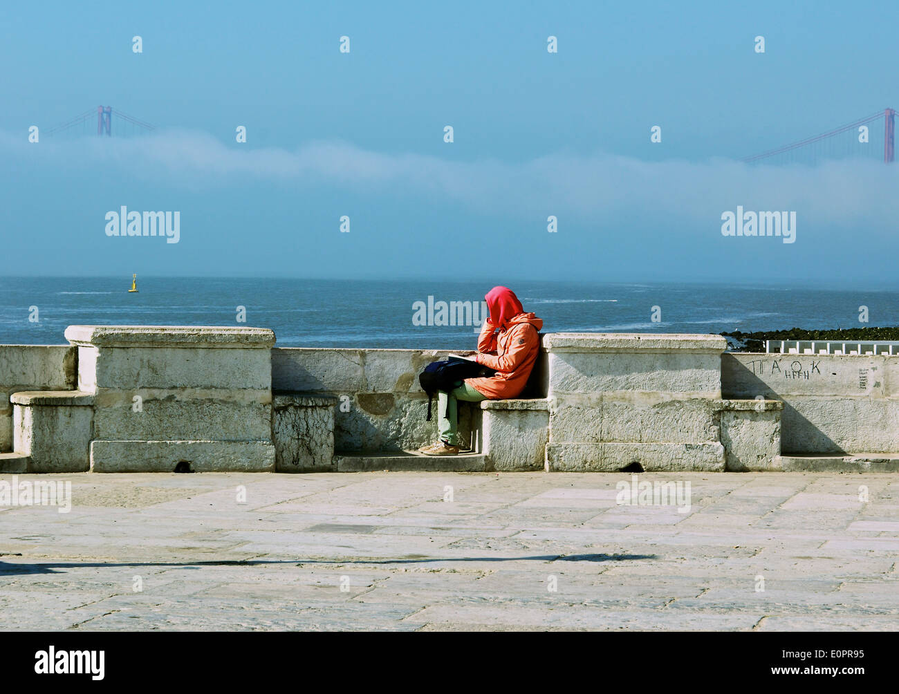Woman sitting reading alone by river Tagus (Tejo) with April 25th bridge in mist Lisbon Portugal western Europe Stock Photo