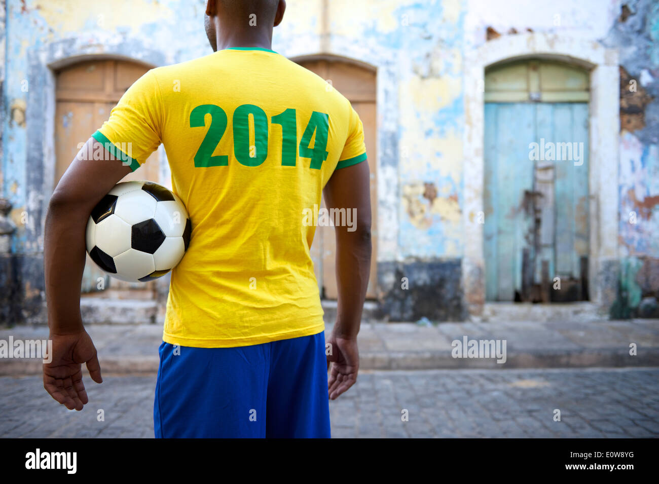 Brazilian football player in 2014 shirt stands holding a soccer ball on an old favela street in rustic village Stock Photo