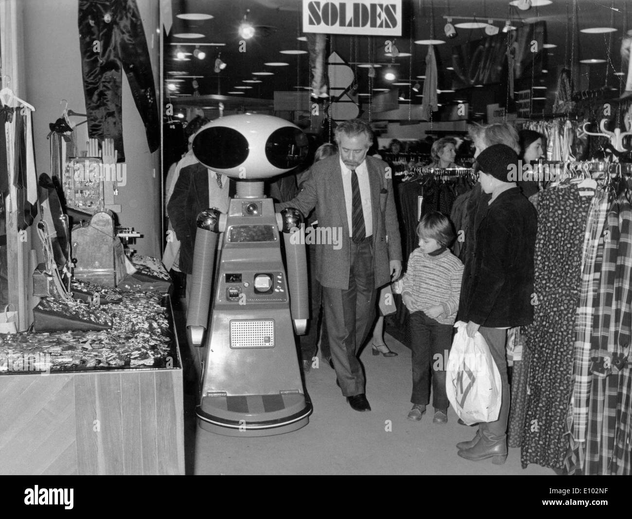 A man walks alongside a alien faced robot inside a French store as a curious woman and two children watch. Stock Photo
