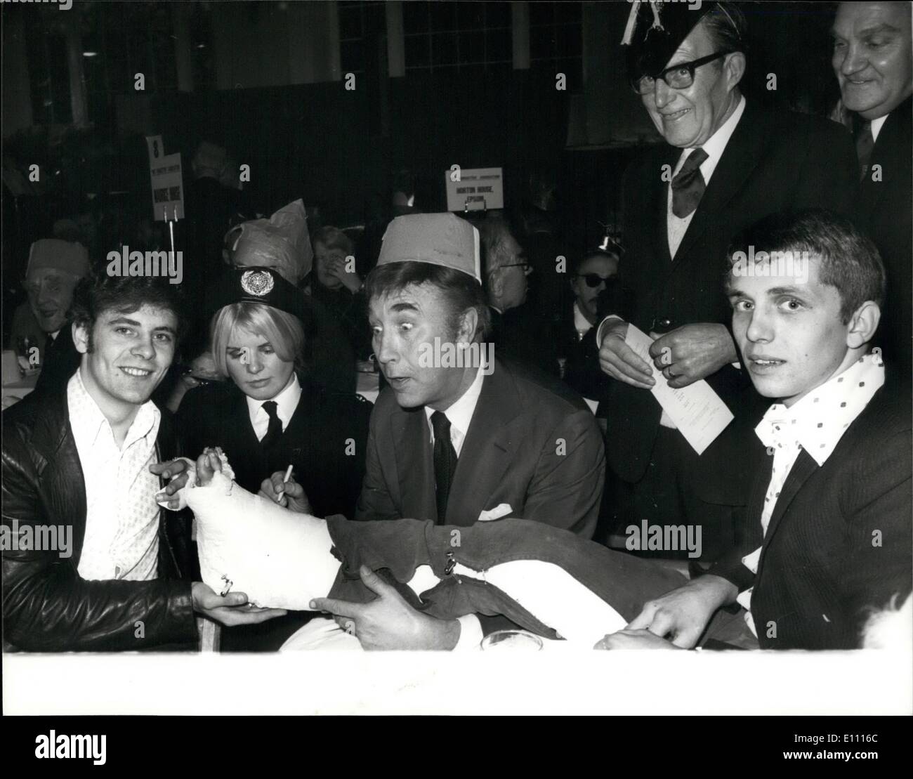 Dec. 12, 1974 - ''Not Forgotten'' Association Christmas Party.: The duchess of Kent, as Patron of the ''Not Forgotten'' Association, this afternoon attended the Association's annual Christmas Party, at which some 450 disabled ex-Service men and women were entertained in the Royal Riding School, Buckingham Palace. Photo shows entertainer Frankie Howerd, pictured at the Christmas Party with two of the victims of the recent public house bomb explosion in Woolwich - who are LT. Col Spellman (seated left), and Gunner Smith, whose plastered leg is being singed by Frankie Howerd. Looking on is St Stock Photo