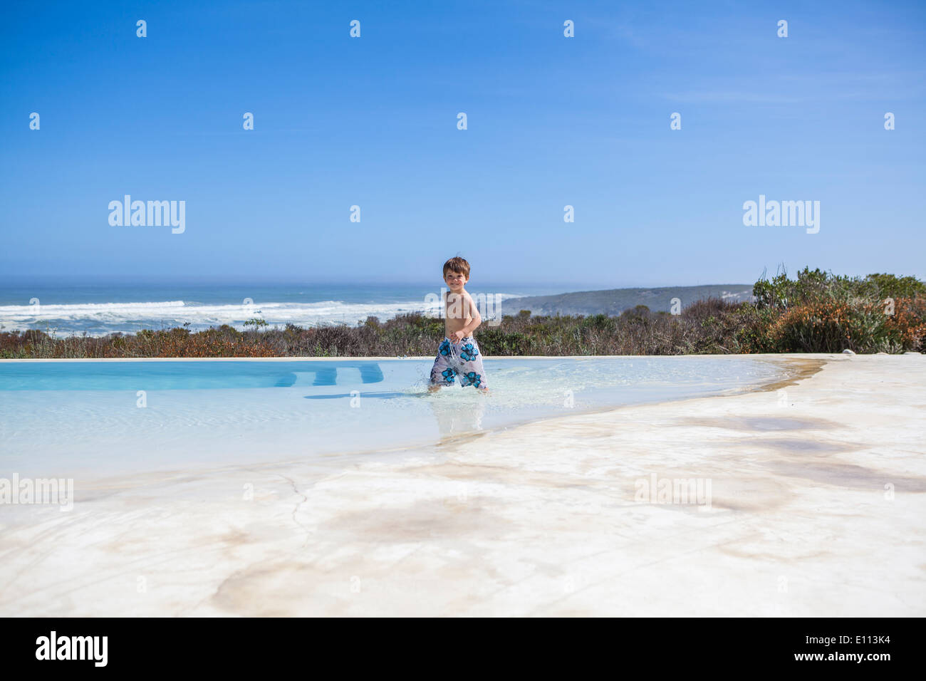Young boy riding a red scooter in board shorts, on a white surface, infinity rim swimming pool and ocean in the background Stock Photo