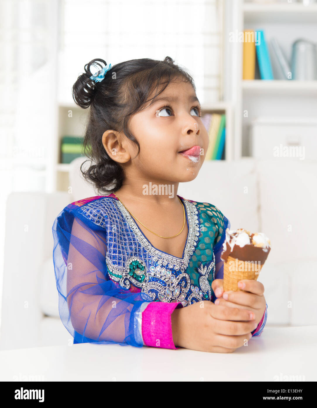 Little Indian girl licking her lips with an ice cream cone in hand, family living lifestyle at home. Stock Photo