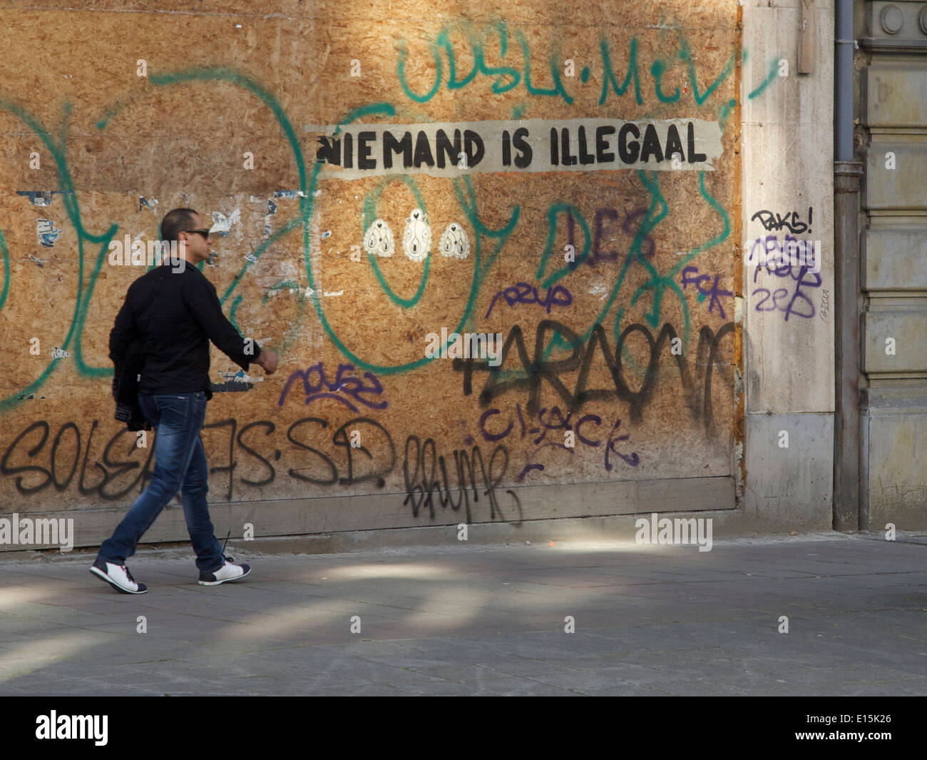 Man walking in a street with graffiti saying nobody is illegal in Dutch