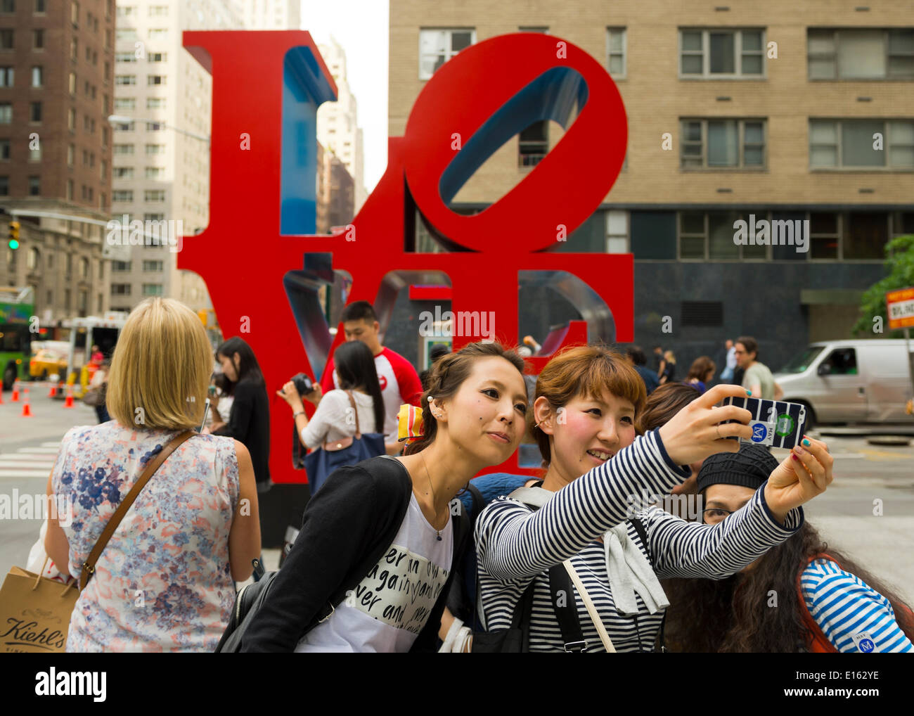 Manhattan, New York, U.S. - May 21, 2014 - Several girl friends take Selfies photos with a cell phone, with the colorful famous LOVE sculpture by Robert Indiana on 6th Avenue in the background, during a pleasant Spring day in Manhattan. Stock Photo
