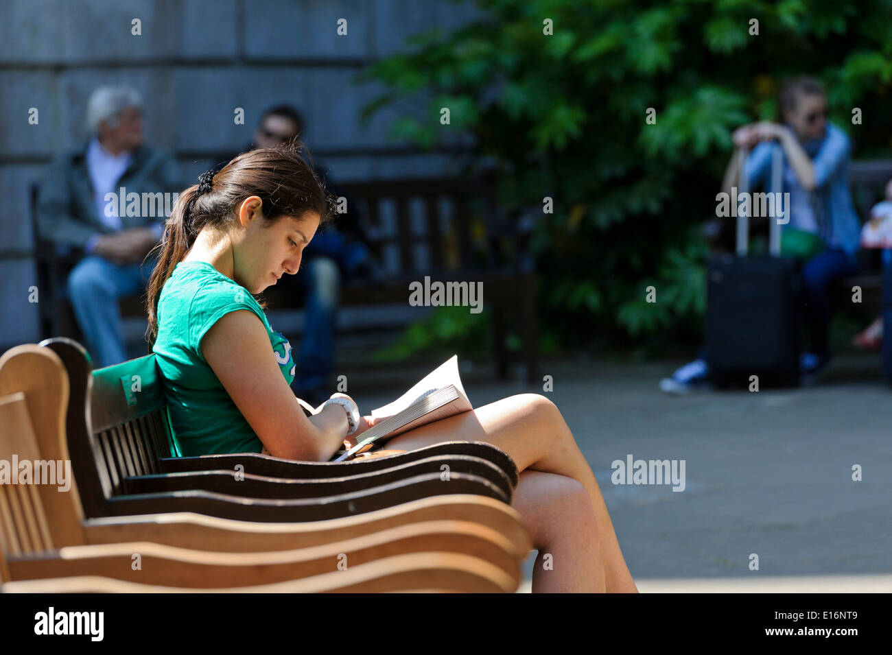 A young woman sitting on a bench reading a book outdoors in a ray of ...