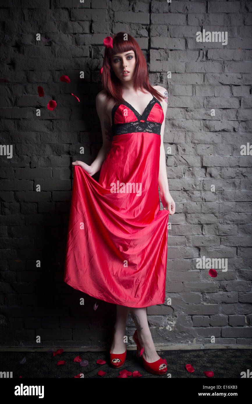 Tattooed woman in a red dress wearing red shoes stands by a wall while rose petals fall upon her. Stock Photo