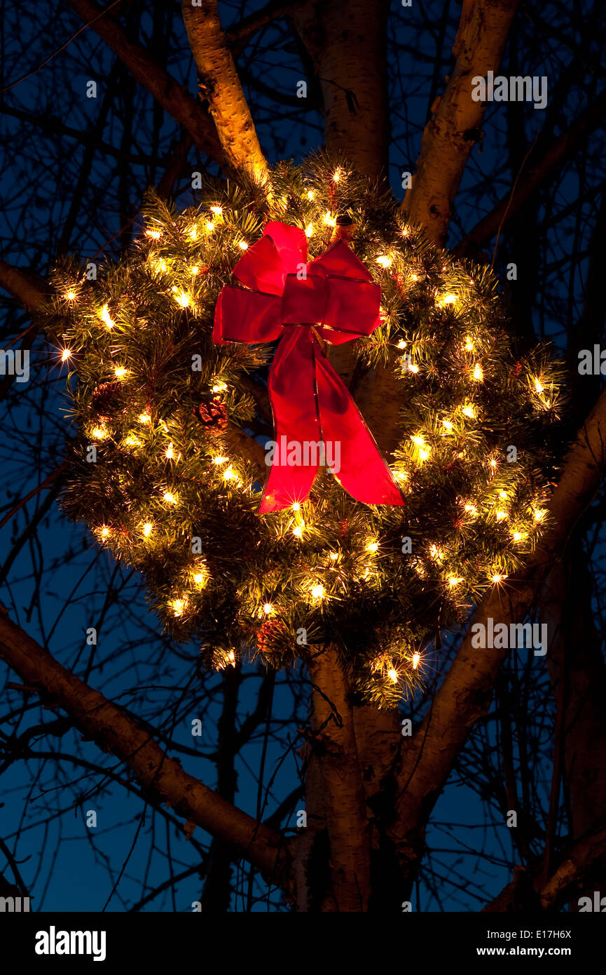 Lit Christmas wreath with red bow hanging in tree during the Holiday season Stock Photo