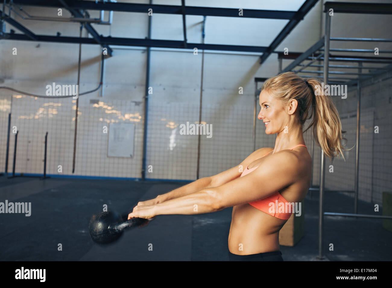Side view of happy young woman exercising with kettle bell at gym. Caucasian female athlete doing crossfit workout at gym. Stock Photo