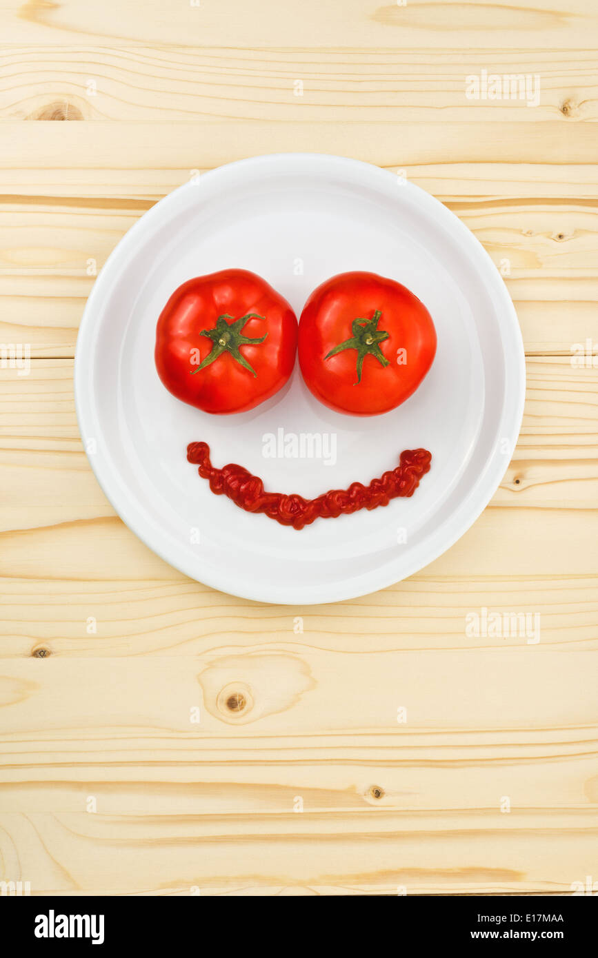 Fun food for children - tomatoes and ketchup making smiley face served on a white plate on wooden table. Stock Photo