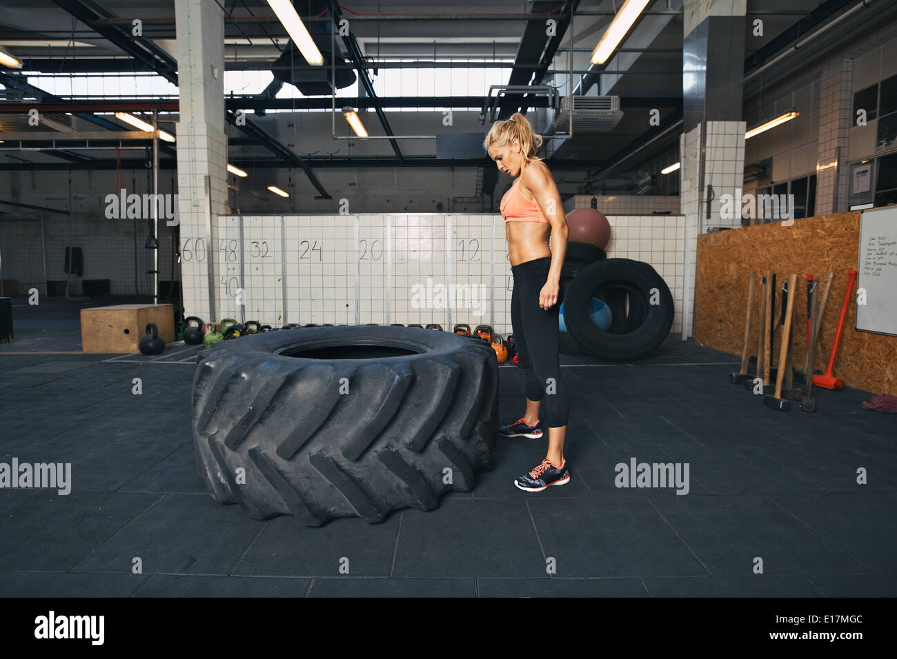 Strong young woman athlete standing and looking at huge tire at gym. Fit female athlete performing tire flipping exercise. Stock Photo