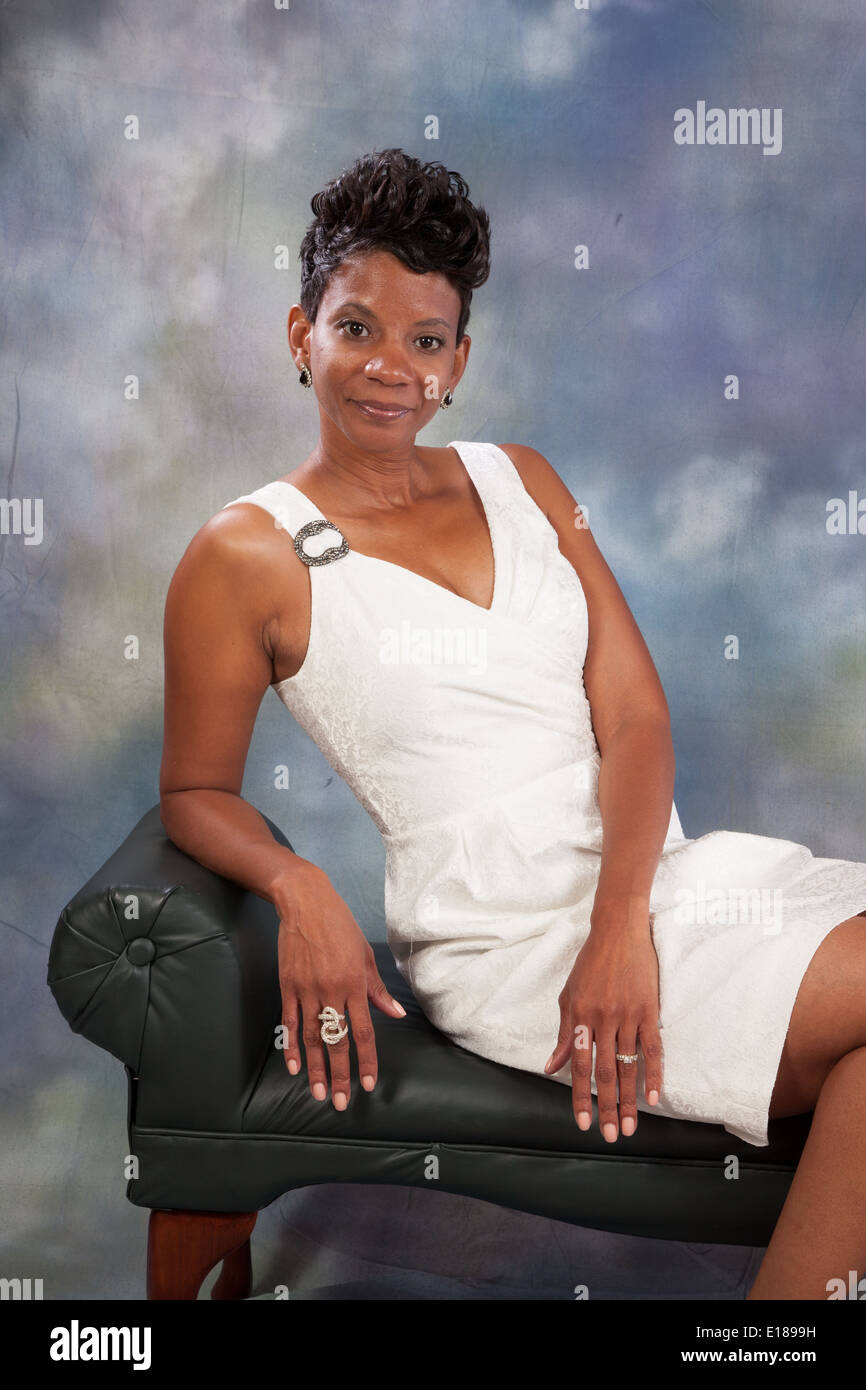 Pretty black woman in white dress, sitting on a bench leaning on an arm, smiling at the camera Stock Photo