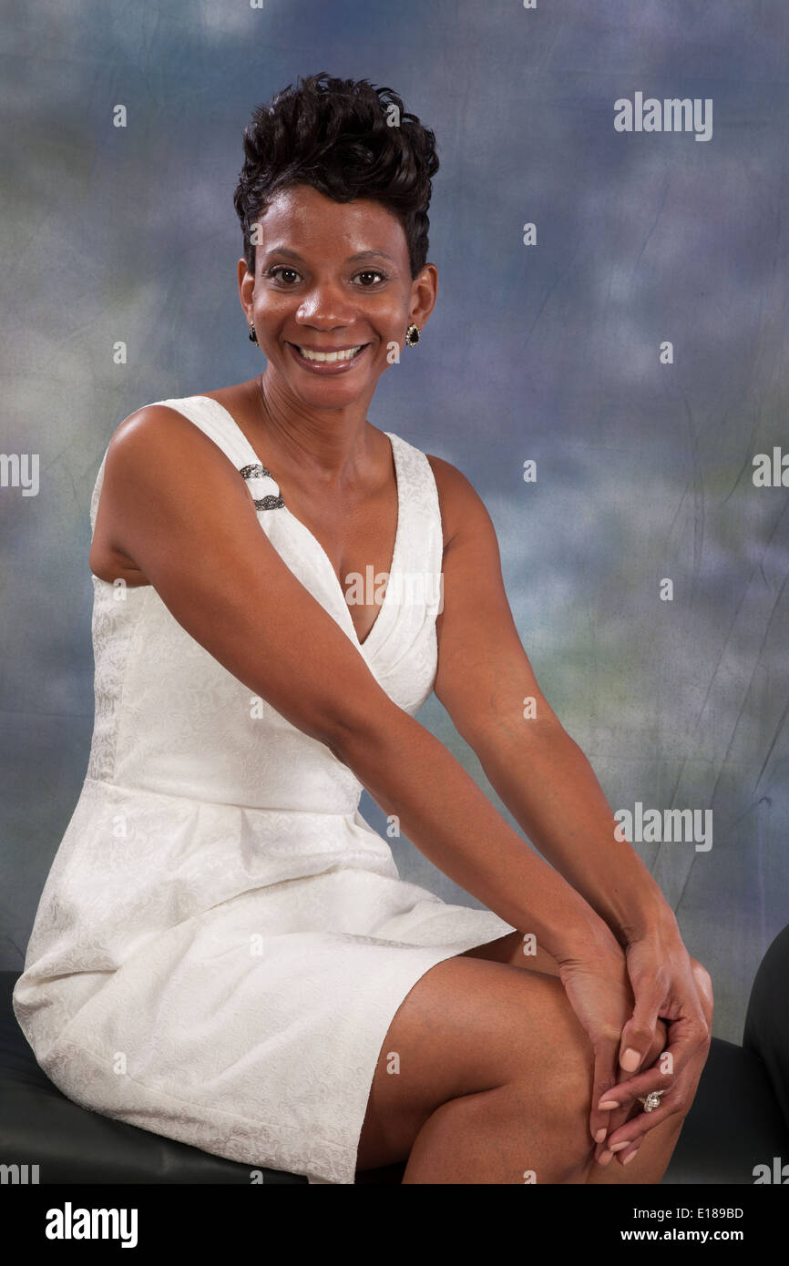 Pretty black woman in white dress, sitting on a bench with her hands on her knees, smiling at the camera Stock Photo