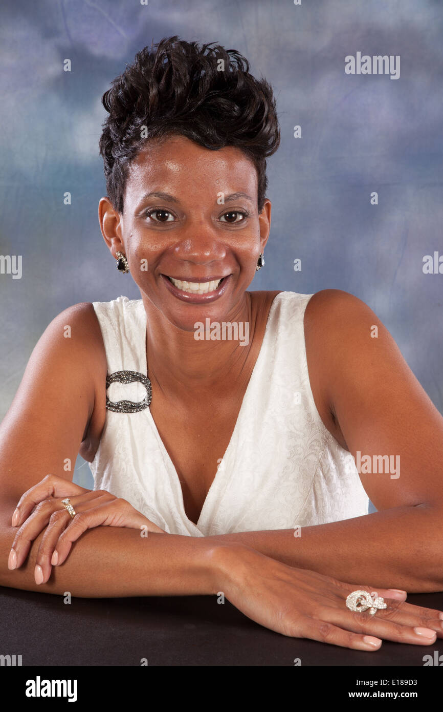 Pretty black woman in white dress, sitting with her arms on a table, smiling at the camera with pleasure and joy Stock Photo