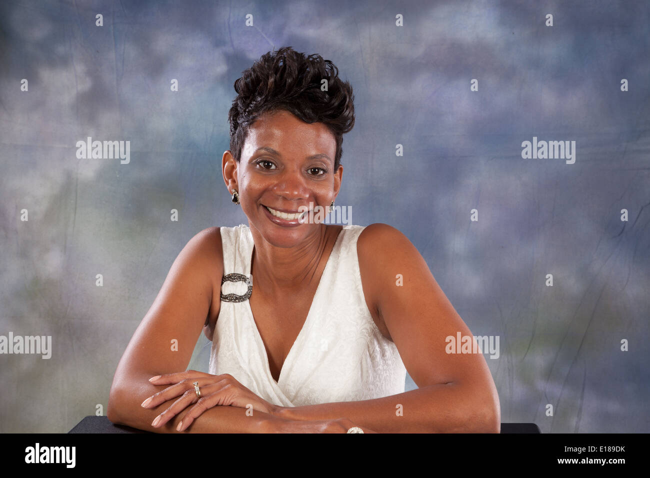 Pretty black woman in white dress, sitting with her arms on a table, smiling at the camera with pleasure and joy Stock Photo