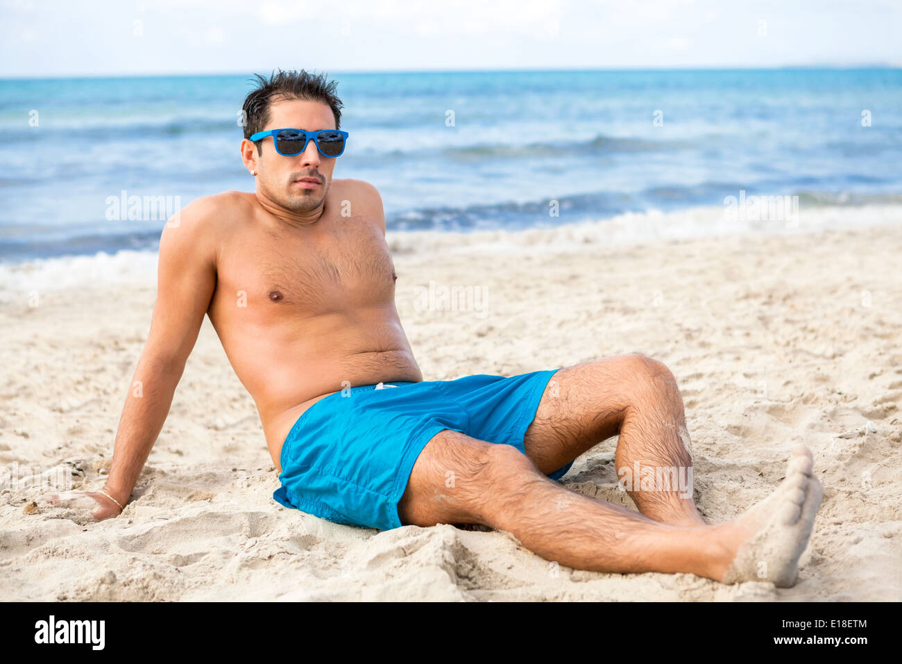 Handsome man wearing trendy sunglasses and his swimsuit relaxing on the  beach sitting on the golden beach sand soaking up the summer sun with the  ocean behind him Stock Photo - Alamy