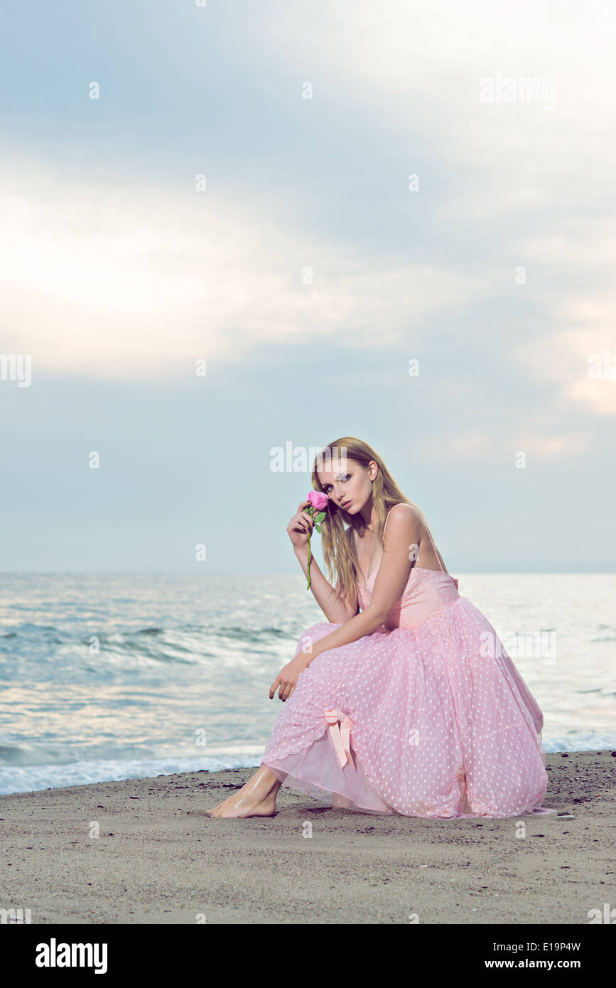 A beautiful young woman portrait sitting on a beach, dressed in a formal pink 50s dress, holding a rose to her face. Stock Photo