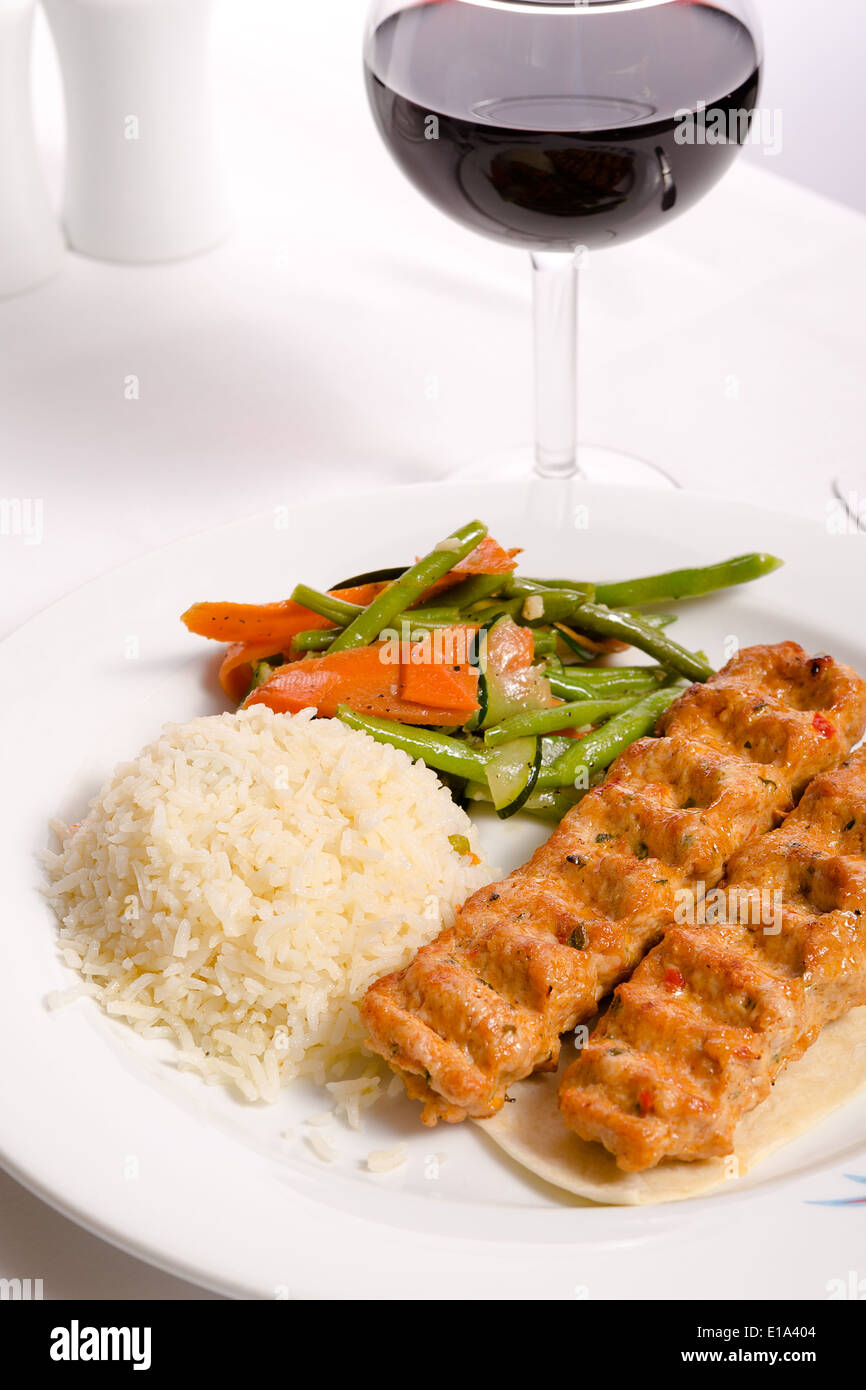 Chicken Adana kebap served with rice pilaf and green vegetables and some carrots, complimented with red Vine and water Stock Photo