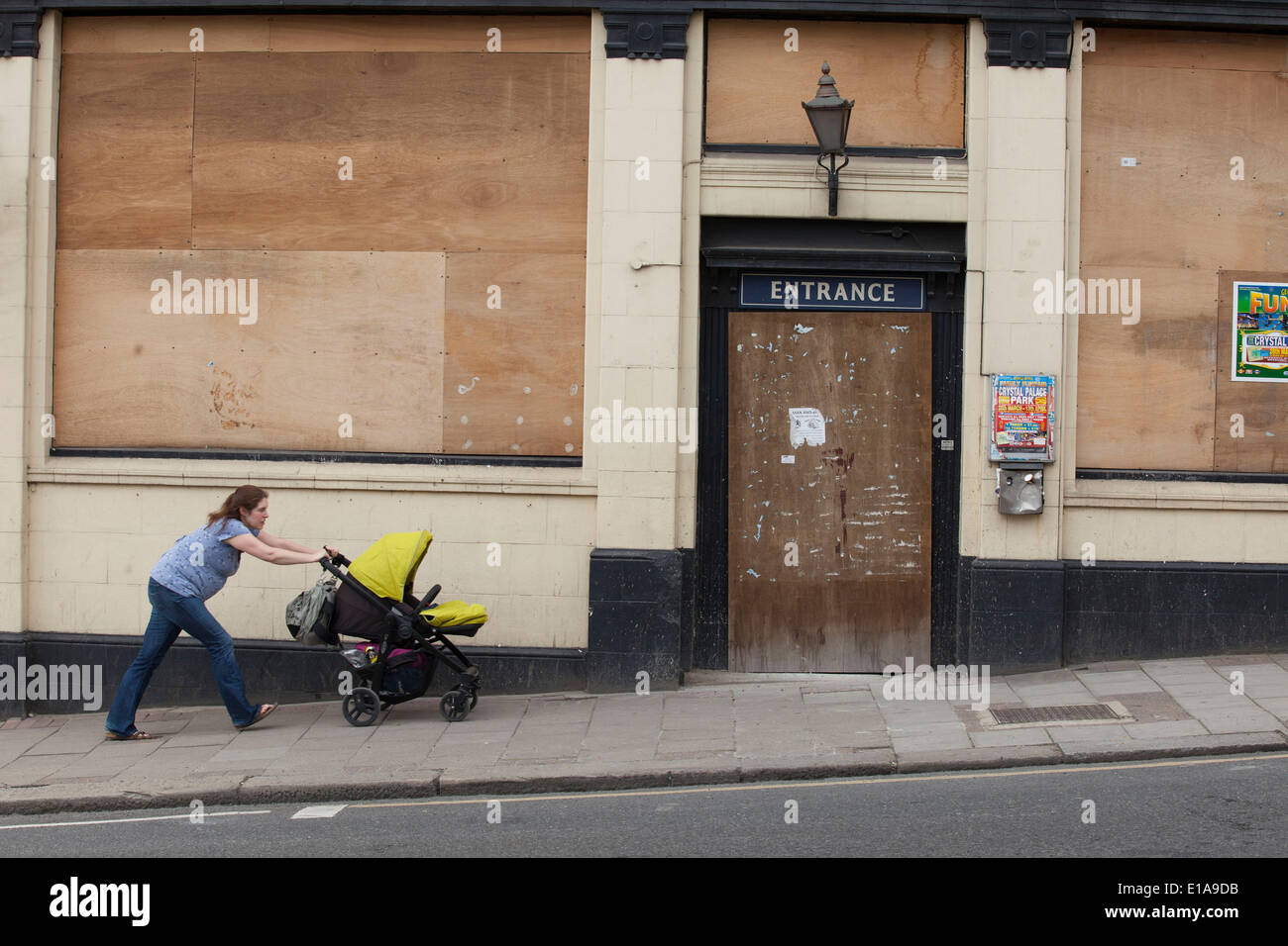Woman pushes a pram past a closed down boarded up pub. Signs of the continued economic downturn and recession in the UK. Stock Photo