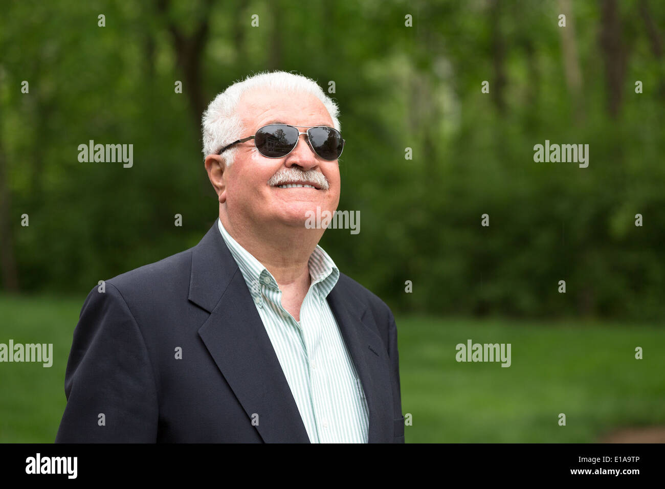 Senior male with grey hair looking up happily and trustfully with his glasses in front of greenery Stock Photo