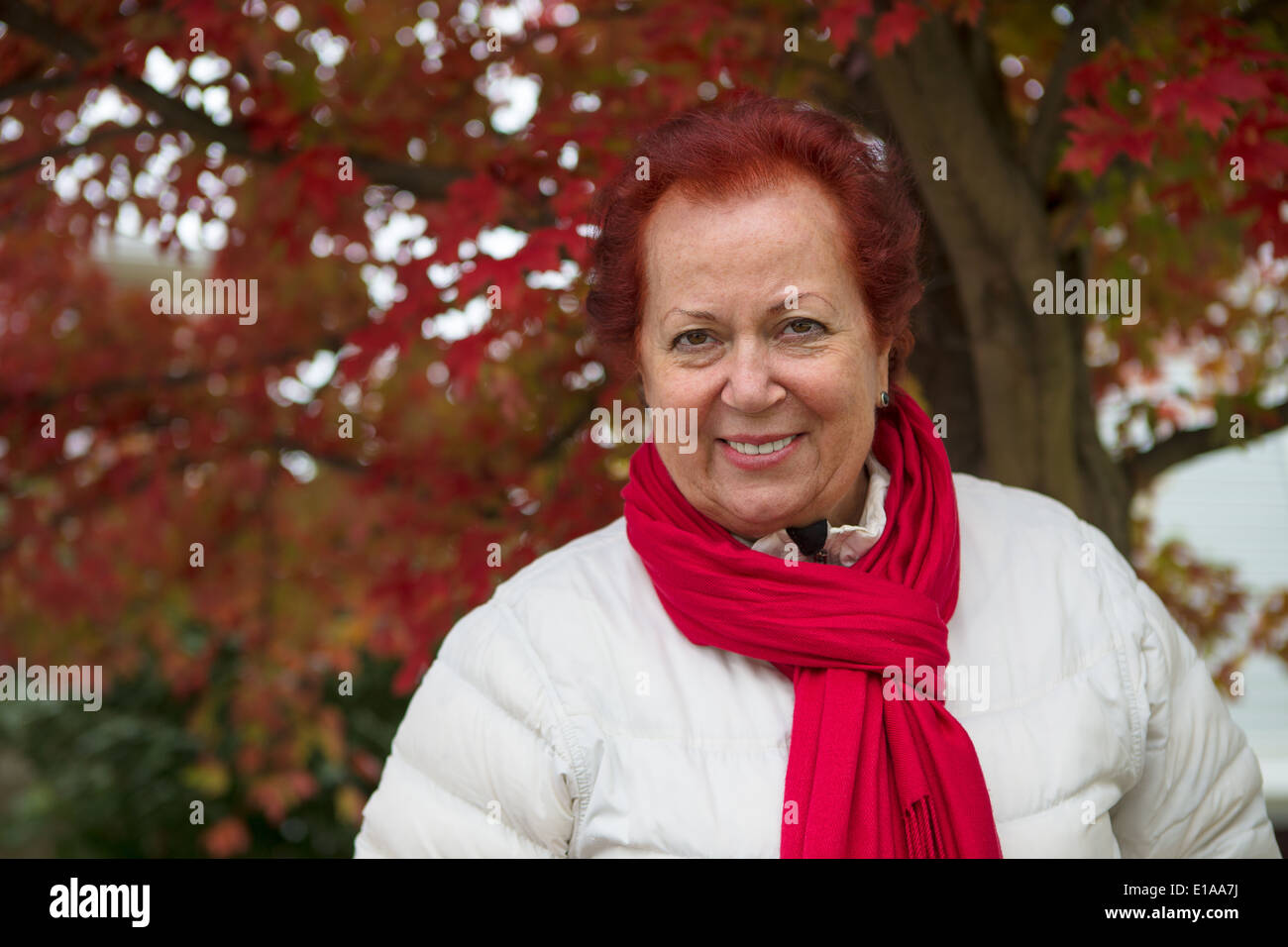 Red hair senior lady looking at you happily and trustfully with her red scarf and white coat under the tree with red falling lea Stock Photo