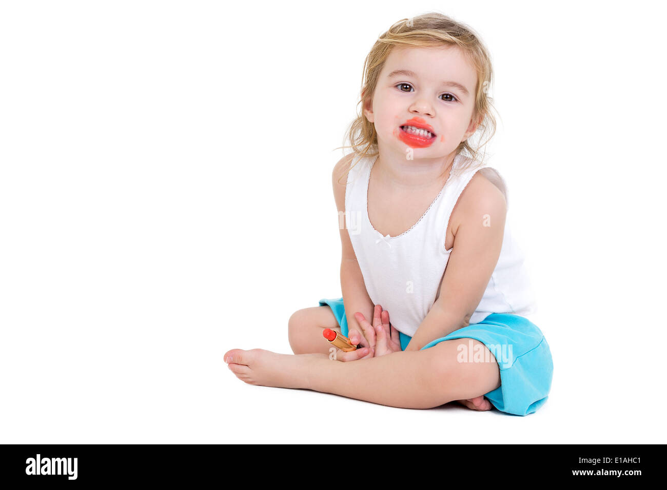 Cute very young girl practising to be a fashion model sitting on the floor grinning at the camera with her mouth smeared with br Stock Photo