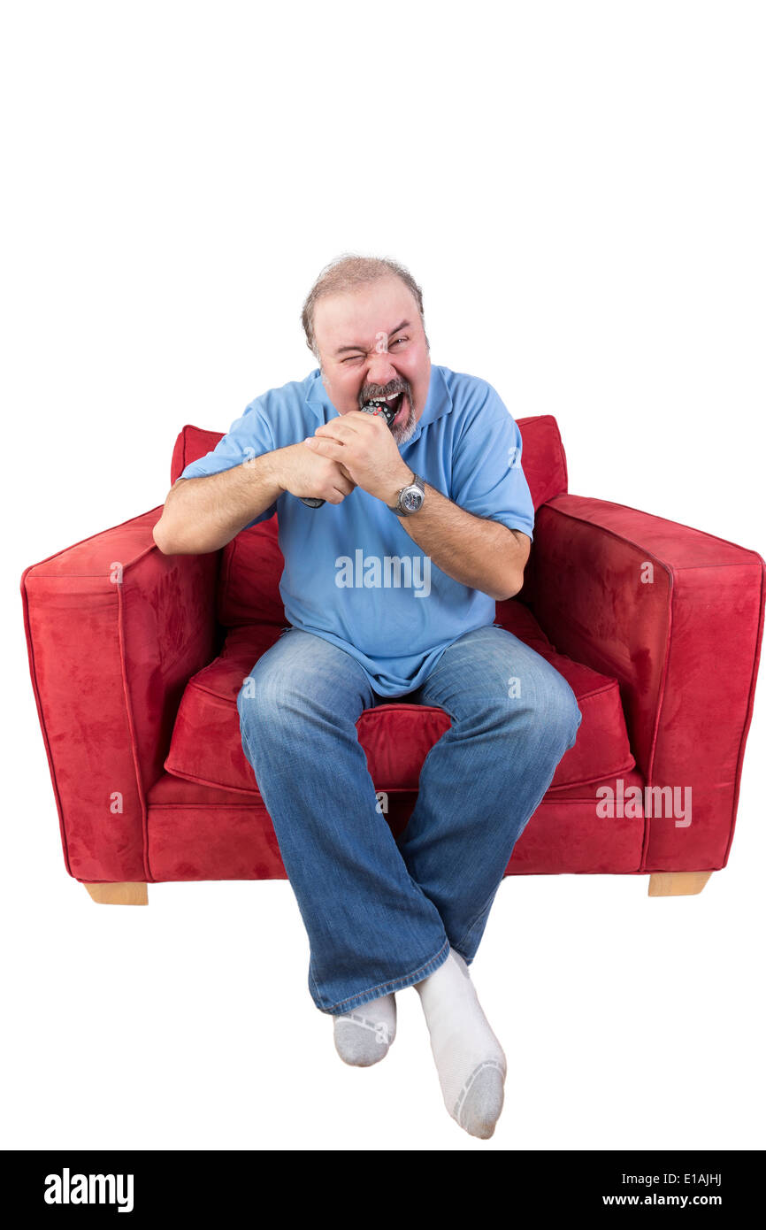 Frustrated middle-aged bearded man sitting in a red armchair biting a remote control from a television set screwing up his eyes Stock Photo