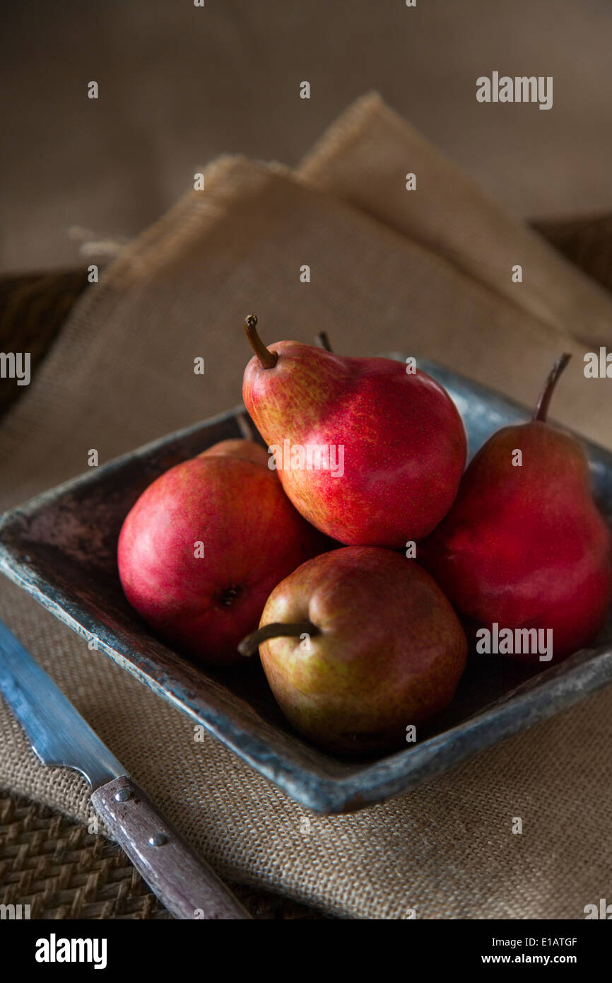 group of red pears in rustic ceramic dish, with knife, on sack cloth and woven tray. Stock Photo