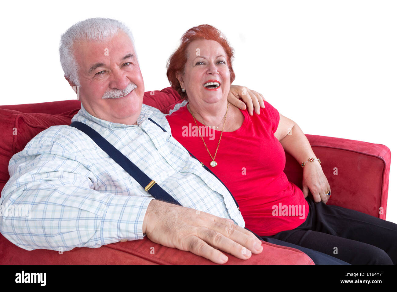 Joyful relaxed elderly couple sitting arm in arm on a red sofa laughing at the camera over white Stock Photo