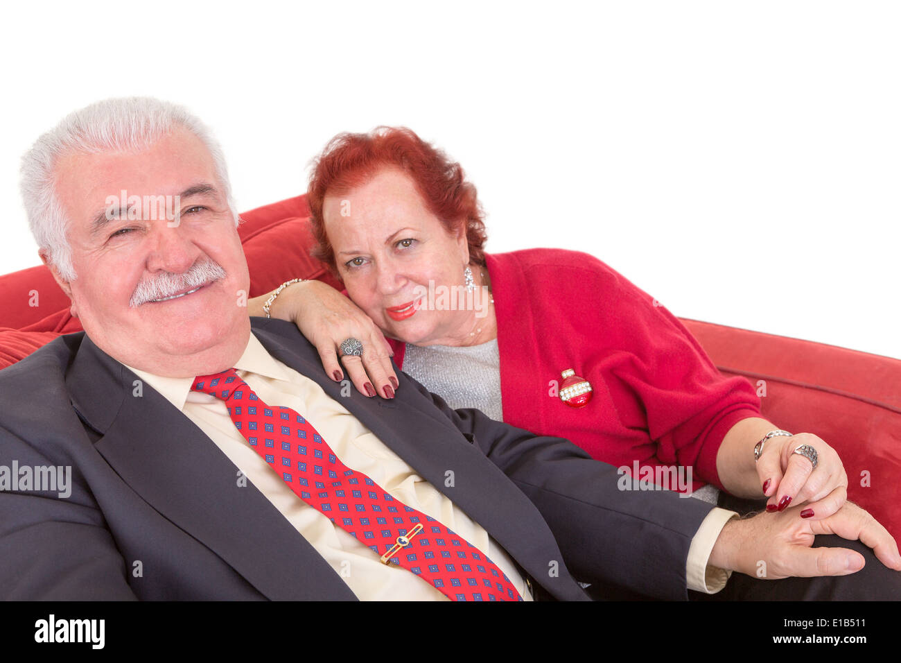 Stylish elderly couple sitting close together side by side on a red sofa looking sideways at the camera with a smile over a whit Stock Photo