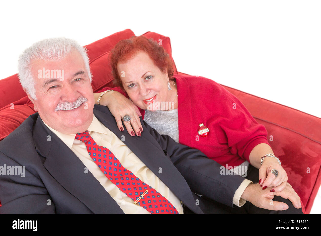 Senior couple sitting on a comfortable red couch holding hands affectionately and looking up at the camera with friendly smiles Stock Photo